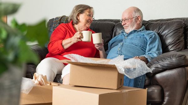Ein älteres Paar sitzt auf einem Sofa vor einem Stapel Kartons und stößt mit Tassen an.