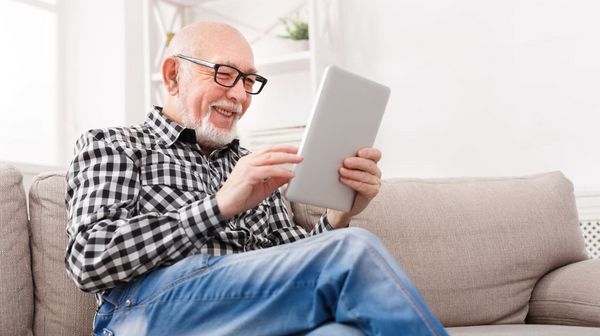 Älterer Mann sitzt mit einem Tablet auf einem beigen Sofa und lacht zufrieden.