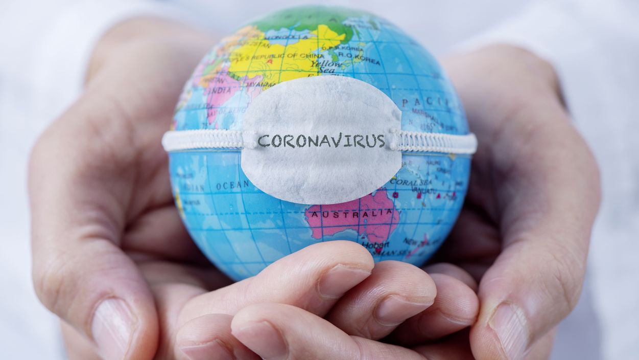 Hände halten eine Weltkugel, um die ein Mund-Nasen-Schutz mit der Aufschrift „Coronavirus“ gespannt ist.